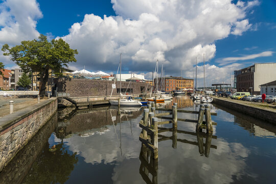 View of shimmering water reflecting the clear sky, puffy clouds, and moored sailboats, framed by brick walls and greenery, Stralsund, Mecklenburg-Vorpommern, Germany.