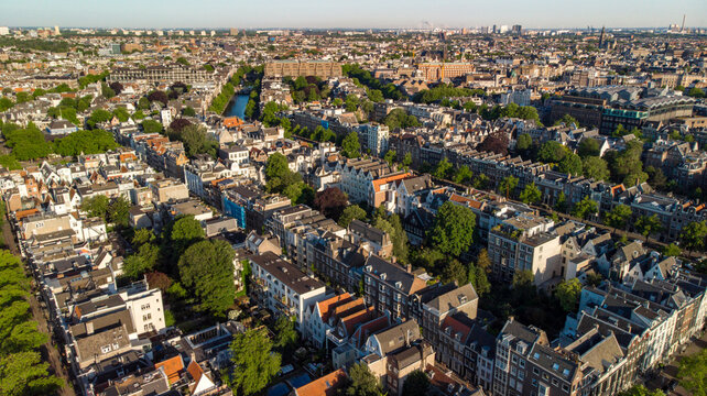 Aerial view of rooftops and canals interspersed with lush green trees reflecting sunlight, creating a mosaic of urban nature, Amsterdam, North Holland, Netherlands.