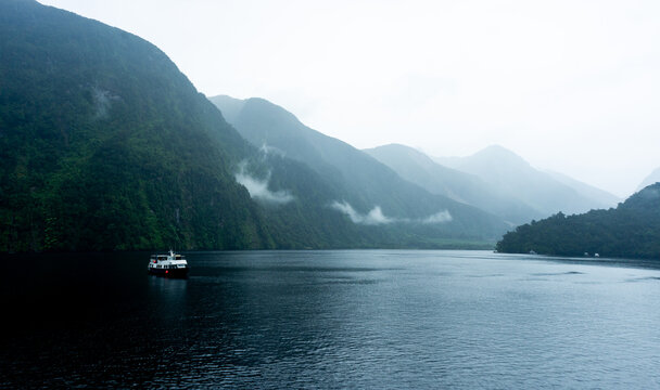 View of a ship sailing through the calm, dark waters surrounded by towering, lush green mountains shrouded in mist, a serene scene, Doubtful Sound, Southland Region, New Zealand.