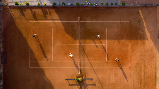 Aerial view of players on a terracotta tennis court, bathed in golden sunlight, casting long shadows across the meticulously lined field, Sint Maarten, North Holland, Netherlands.