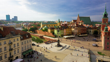 View of a vibrant city square bustling with life, framed by colorful buildings and a historic castle under a bright sky, Warsaw, Masovian Voivodeship, Poland.