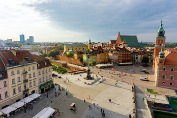 View of a vibrant city square bustling with people, surrounded by colorful buildings and historic architecture under a clear sky, Warsaw, Masovian Voivodeship, Poland.
