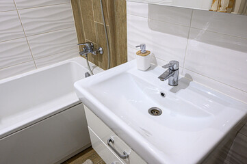 modern bathroom features a white ceramic sink with a chrome faucet, a soap dispenser, a white bathtub with a chrome shower tap, light-toned tiles, and wooden-style wall panels