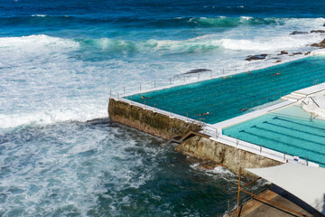View of azure waters collide against the concrete edges of Bondi's iconic pool, where swimmers glide amidst the crashing waves, Bondi Beach, New South Wales, Australia.