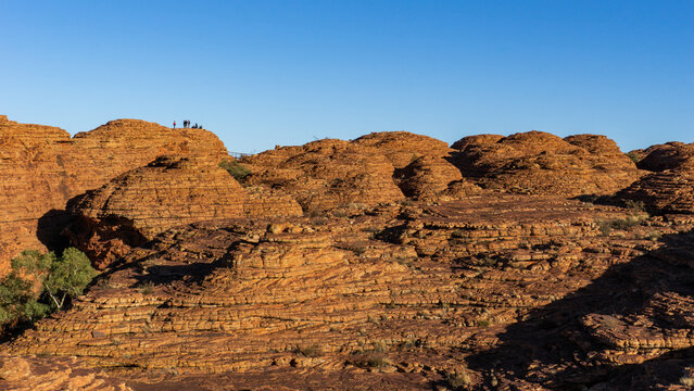 View of the rugged, textured sandstone formations under a clear blue sky, with tiny figures standing atop, Kings Canyon, Northern Territory, Australia.