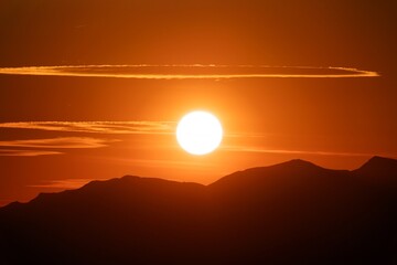 A vibrant orange sunset casting a warm glow over dark mountain silhouettes with a unique circular cloud formation above the sun.