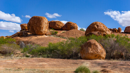 View of colossal, weathered boulders scatter across the ochre landscape under a vast blue sky dotted with fluffy clouds, Devils Marbles, Warumungu, Northern Territory, Australia.