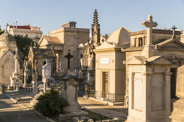 View of ornate mausoleums and crosses stand in solemn silence under the warm sun, with buildings in the background, Barcelona, Catalonia, Spain.