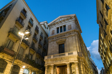 View of golden light highlighting the ornate facades of historic buildings under a twilight sky, framing a classic European streetscape, Barcelona, Catalonia, Spain.