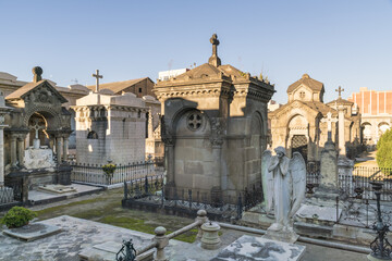 View of aged mausoleums and gravestones with weathered stone angels stand in solemn silence under a clear sky, Barcelona, Catalonia, Spain.