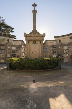 View of a somber, symmetrical mausoleum capped with a stone cross, surrounded by trimmed green shrubs in a sunlit cemetery, Barcelona, Catalonia, Spain.