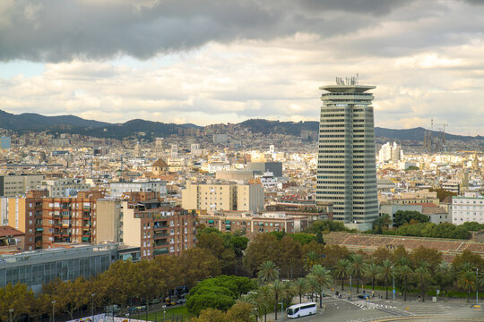 View of a modern Torre Mapfre tower rising above the terracotta rooftops and green trees under a cloudy sky, Barcelona, Catalonia, Spain.
