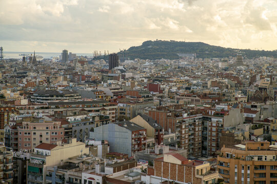 View of rooftops sprawl under a soft, diffused sunlight, blending with the distant, hazy silhouette of mountains and the glint of the sea, Barcelona, Catalonia, Spain.