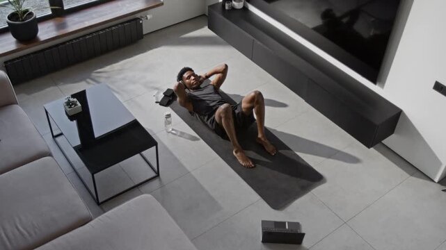 High angle view of Black young sportsman with headphones doing crunches on yoga mat during home workout with laptop, fitness bands and water bottle placed nearby