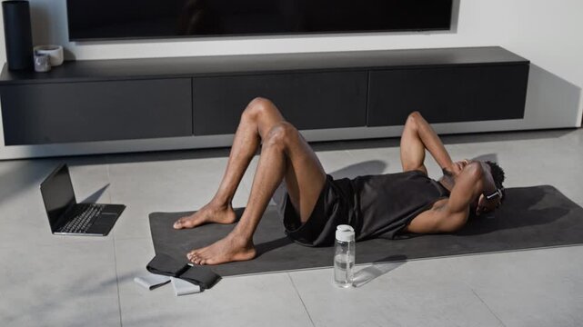 Black man performing abdominal crunches on exercise mat in modern room, with open laptop, resistance bands and and fitness bottle placed on floor beside him