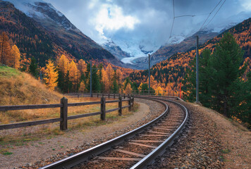 Curved railway tracks through autumn forest in the Swiss Alps with snow-covered mountains in clouds and glacier. Mountain railroad infrastructure in alpine landscape, Switzerland. Railway station