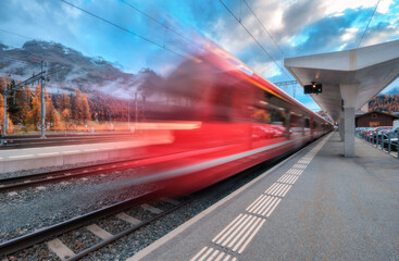 Blurred red passenger train passing mountain railway station in Swiss Alps at dusk. Moving high-speed train. St. Moritz, Switzerland. Bernina Express. Railway platform and cloudy sky. Rail transport