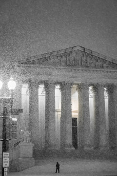View of a solitary figure braving the blizzard's wrath before the majestic columns of a neoclassical building, bathed in eerie light, Washington DC, United States.