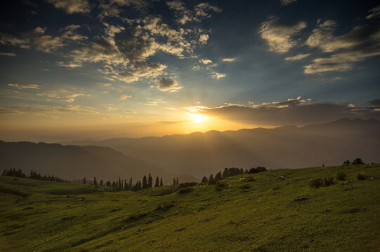 View of a golden sunset casting long shadows over rolling green hills and distant mountains, Sri Paye, Khyber Pakhtunkhwa, Pakistan.