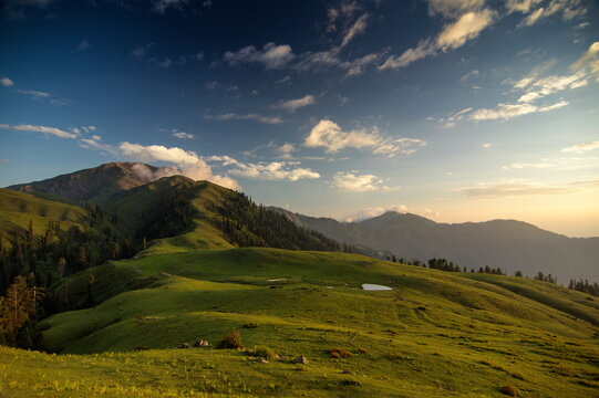 View of sunlight kissing the verdant hills and distant mountain peaks under a blue sky dotted with soft clouds, Sri Paye, Khyber Pakhtunkhwa, Pakistan.