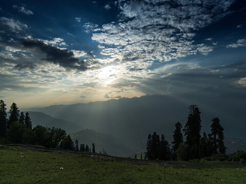 View of sunbeams pierce through a dramatic sky, illuminating the rolling hills and lush greenery in the distance, Sri Paye, Khyber Pakhtunkhwa, Pakistan.