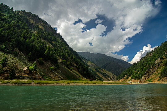View of emerald waters reflecting the sky beneath towering, evergreen-clad mountains under a sky brushed with white clouds, Sri Paye, Khyber Pakhtunkhwa, Pakistan.