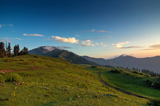 View of lush green hills roll under a bright sky towards distant, misty mountains, capturing the serene beauty of nature, Sri Paye, Khyber Pakhtunkhwa, Pakistan.