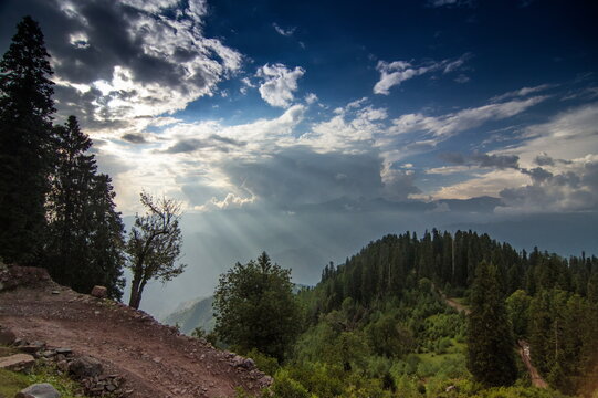 View of sunlight piercing through dramatic clouds over lush green hills and dense forests creating a serene landscape, Sri Paye, Khyber Pakhtunkhwa, Pakistan.