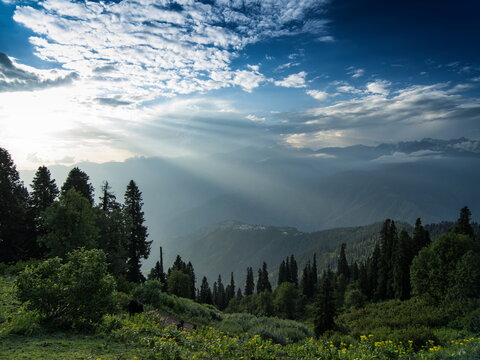 View of sun rays piercing through dramatic clouds over a verdant mountain landscape filled with trees, Sri Paye, Khyber Pakhtunkhwa, Pakistan.