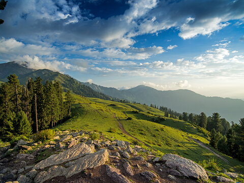 View of lush green hills rolling under a vibrant blue sky dotted with fluffy clouds, framed by rugged rocks in Sri Paye, Khyber Pakhtunkhwa, Pakistan.