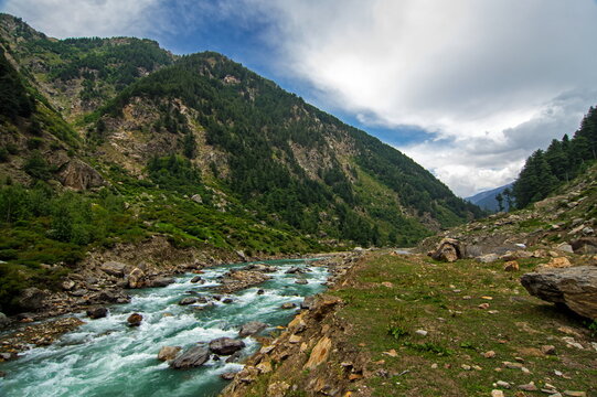 View of turquoise river rushing through a rugged valley, flanked by steep, rocky slopes and dense green forests under a cloudy sky, Sri Paye, Khyber Pakhtunkhwa, Pakistan.