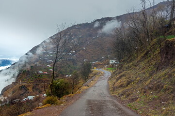 View of a winding asphalt road cutting through a steep mountainside dotted with houses, shrouded in mist and clouds, Pir Chinasi, Azad Kashmir, Pakistan.