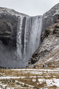 View of powerful Skogafoss waterfall cascading down rugged cliffs dusted with snow, while tiny figures explore the landscape below, Iceland.