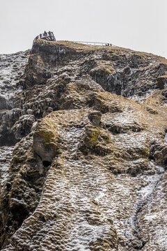 View of rugged, snow-dusted cliffs rise against a muted sky, with tiny figures silhouetted atop the highest peak, a cold, wild beauty unfolds, Skogafoss, Iceland.