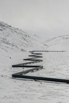 View of a bold, dark pipeline snakes across a snow-dusted landscape, carving a path through the white expanse under a muted sky, Iceland.