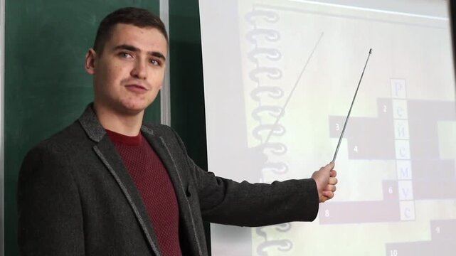 Young male teacher in jacket pointing with stick at projected slide or screen in classroom. Educational presentation. The word rheismus in Ukrainian