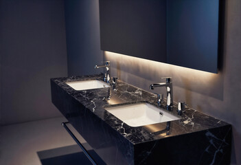 Modern black marble bathroom vanity with dual sinks and chrome faucets under a backlit mirror.