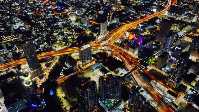 Downtown Miami at night, with cars driving on illuminated roads in a urban skyline scene. Aerial view. Street and house lighting. River with a boat.
