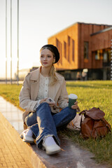 A young girl sits on a green lawn in a park with a cup of takeaway coffee. Lunch break from school or work outside.