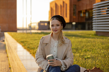 A young girl sits on a green lawn in a park with a cup of takeaway coffee. Lunch break from school or work outside.