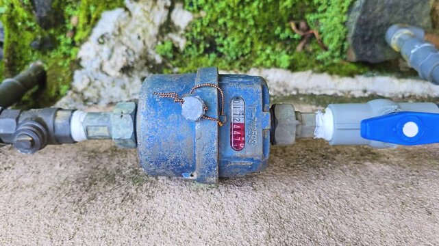 Pull-out shot of a running water meter in a moss-covered abandoned place, with the meter numbers spinning as water flows through aged utility infrastructure. 4K