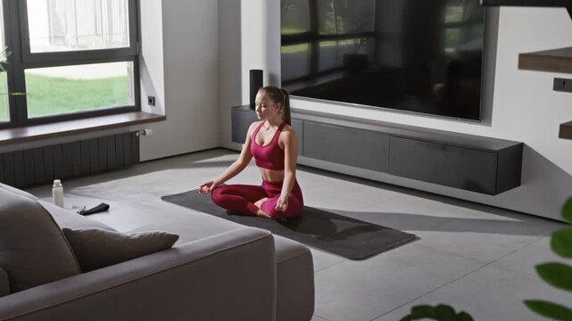 Young sportswoman in activewear meditating with crossed legs on yoga mat during morning practice in modern living room