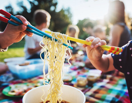 A child uses colorful chopsticks to lift wavy noodles during a sunny park picnic. This vibrant shot evokes the joy of outdoor dining and family bonding. Ideal for lifestyle and food media. - Powered by Adobe