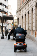 Obraz premium Senior man in a wheelchair scooter navigating a city street, showcasing urban mobility and independence, with modern architecture and pedestrians in the background