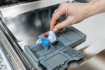 Hand placing colorful dishwasher detergent pods into the compartment of a modern dishwasher, showcasing the cleaning process and efficient home appliance usage with copy space