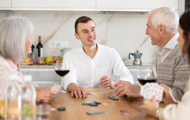 Merry old man and woman and young couple engaged in poker while having red wine in comfort of their house