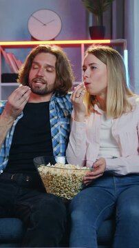 Leisure concept where attractive smiling carefree man and woman resting on comfortable sofa in sitting room at night previsioning interesting show on TV set,tasting popcorn and enjoying conversation