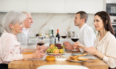 Positive daughter-in-law and mother-in-law exchanging smiles and friendly conversation at dinner table in cozy kitchen, husbands sitting nearby..