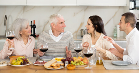 Merry old family members having lunch with their son and his girlfriend while drinking wine and talking warmly in their house