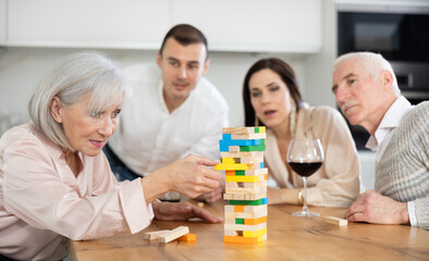 Fototapeta premium Cheerful senior woman spending free time with husband and adult children at laid-back family home get-together with drinks, concentrating on removing block from tower while playing jenga